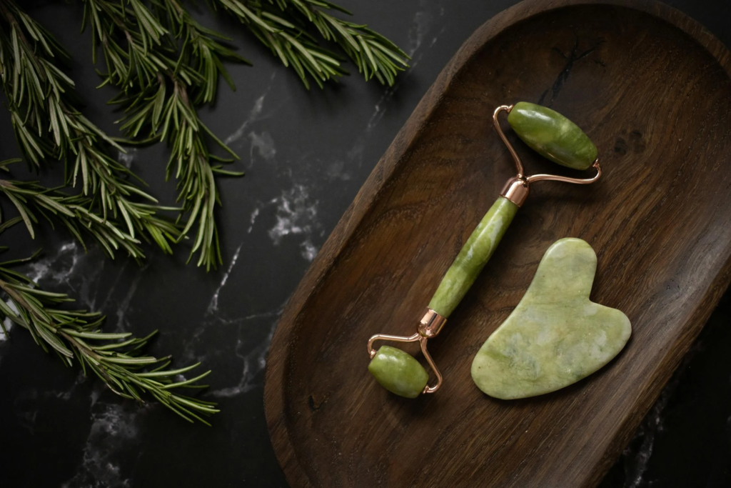 Flat-lay image of a jade roller and Gua Sha stone on a wooden tray, surrounded by fresh sprigs of rosemary, used for facial massage and lymphatic drainage to reduce puffiness and promote skin health.