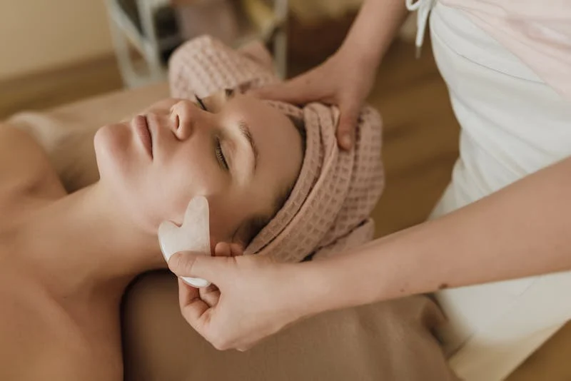 "Esthetician performing a professional Gua Sha facial massage on a woman in a spa setting, using a white Gua Sha stone to promote lymphatic drainage and relaxation.